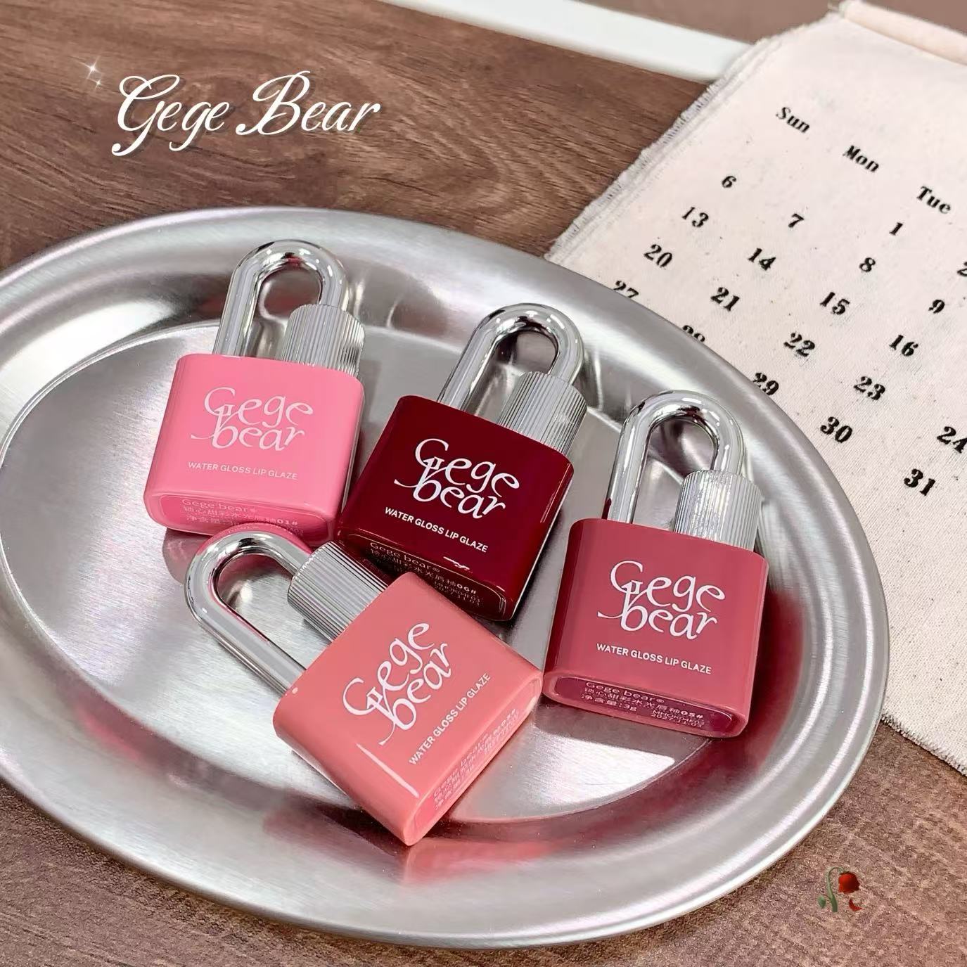 Four small bottles of 'Gege Bear' lip gloss on a metallic tray with a calendar in the background.
